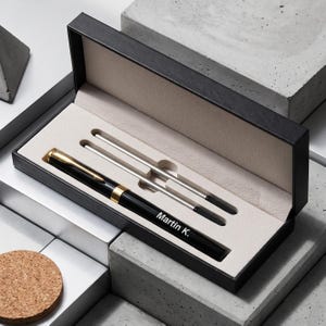 May include: A black fountain pen with gold-tone accents and the name Martin K. engraved on the barrel, presented in a black rectangular gift box with a beige interior lining. The set includes two silver-toned ink refills placed in the box.