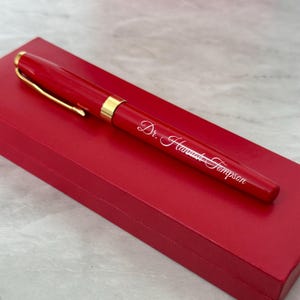 May include: A red pen with a gold trim and the name "Dr.  Hawkins Thompson" engraved on the side. The pen is in a red box.