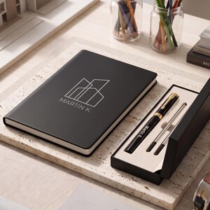 May include: A black notebook with a white geometric building logo and the text MARTIN K. printed on the cover, accompanied by a matching black pen set in a rectangular black gift box with a cream-colored interior tray and gold accents.