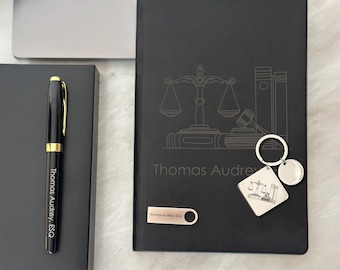 May include: Black leather journal with a silver engraved design of a scale of justice, a gavel, and books. The journal has the name "Thomas Audrey" and the title "ESQ" engraved on the front. A silver keychain with a similar design is included.