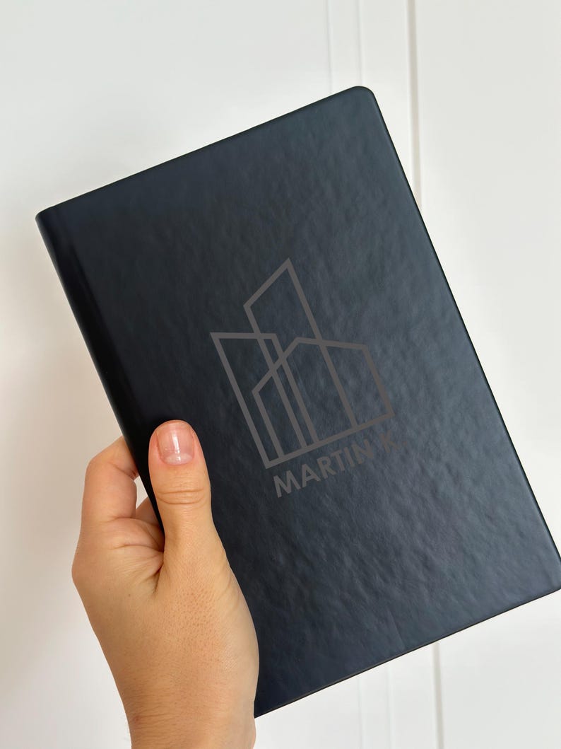 May include: A black rectangular notebook with a textured cover featuring a grey line-art graphic of three overlapping buildings and the text MARTIN K printed in a clean sans-serif font centered on the front cover.