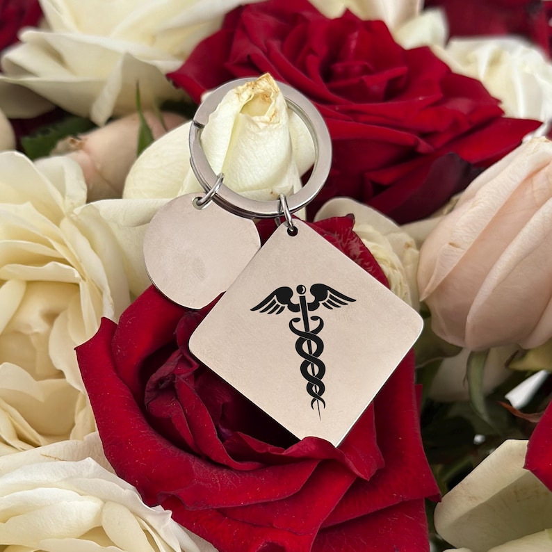 May include: A silver keychain with a round charm and a square charm. The square charm has a medical symbol engraved on it. The keychain is surrounded by white and red roses.