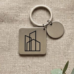 May include: A silver-toned metal keychain featuring a square pendant with a black line-art graphic of three overlapping buildings. The keychain includes a circular split ring and a small, plain, round silver-toned metal charm attached to the ring.