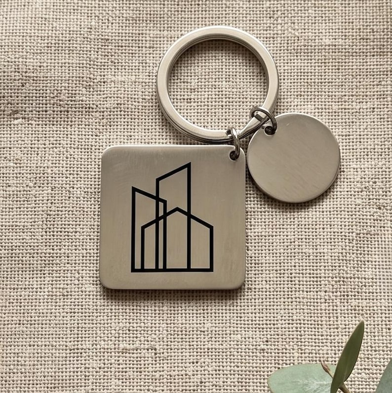 May include: A silver-toned metal keychain featuring a square pendant with a black line-art graphic of three overlapping buildings. The keychain includes a circular split ring and a small, plain, round silver-toned metal charm attached to the ring.