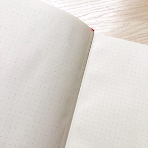 May include: An open notebook with white pages and a dotted grid pattern. The notebook is bound with a red spine.