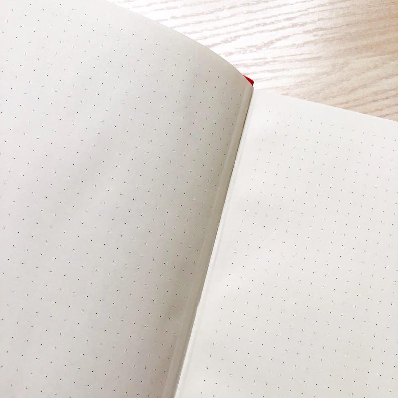 May include: An open notebook with white pages and a dotted grid pattern. The notebook is bound with a red spine.