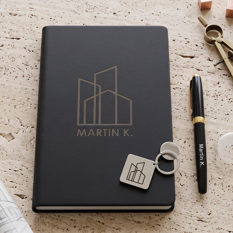 May include: A black notebook featuring a geometric building logo and the text "MARTIN K." embossed on the cover. A matching black pen with the name "Martin K." printed on the barrel and a silver square keychain with the same logo are included.