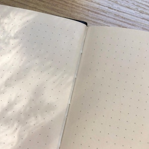 May include: An open notebook with cream-colored pages featuring a black dot grid pattern. The pages are bound together at the center spine, showing a clear fold where the two sides meet. The paper has a smooth texture and consistent dot spacing.