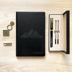 Op de afbeelding: Een zwart lederen notitieboek met een bergketenontwerp en de naam "V. Roberts" gegraveerd op de voorkant. Het notitieboek wordt geleverd met een zwarte pen en een zilveren sleutelhanger met een sleutelring en een kleine zilveren hanger.