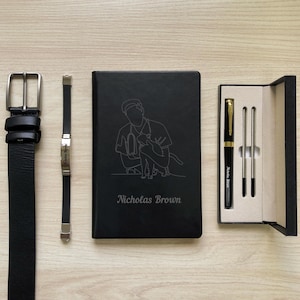 Puede incluir: Un cinturón de cuero negro, una pulsera plateada, una libreta negra con un dibujo de línea de una persona sosteniendo un gato, el texto "Nicholas Brown", y un juego de bolígrafo y lápiz negro en una caja negra.