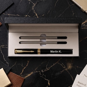 May include: A black fountain pen with gold-tone accents and the name Martin K. engraved on the barrel, presented in a black rectangular gift box with a cream-colored interior. The set includes two additional ink refills stored in the box.