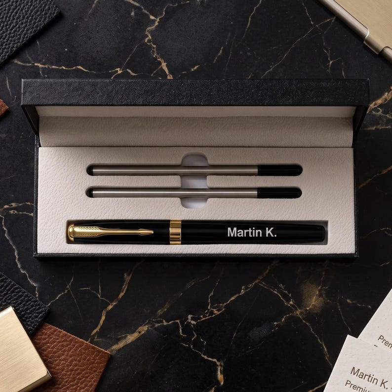 May include: A black fountain pen with gold-tone accents and the name Martin K. engraved on the barrel, presented in a black rectangular gift box with a cream-colored interior. The set includes two additional ink refills stored in the box.