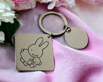 May include: A silver keychain with a square charm featuring a black and white illustration of a bunny holding a teddy bear. The keychain also has a round silver tag.