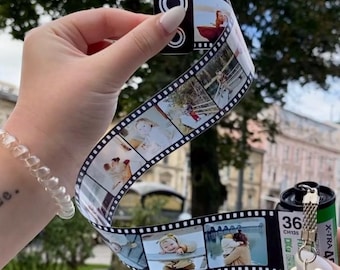 Op de afbeelding: Een sleutelhanger met filmstrip met een reeks kleine foto's. De filmstrip wordt door een persoon vastgehouden. De sleutelhanger bevat een filmcassette met het nummer 36 erop. De filmstrip toont een reeks foto's.