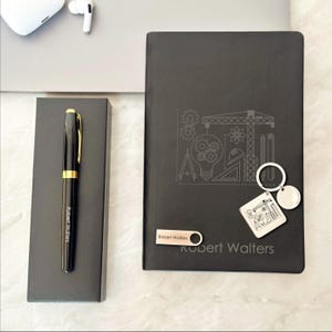 Könnte beinhalten: Ein schwarzer Stift mit goldenen Akzenten und eine passende Box, ein schwarzes Notizbuch mit einem technischen Design, ein Schlüsselanhänger und ein USB-Stick. Auf dem Notizbuch und der Stiftbox steht der Name "Robert Walters".