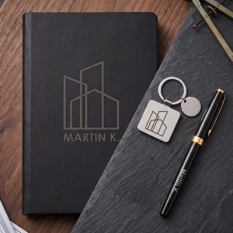 May include: A black notebook with a building logo and the text MARTIN K. in gold, a square silver keychain with the same building logo, and a black pen with gold accents featuring the text Martin K. engraved on the side of the barrel.