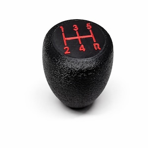 Peut inclure: Pommeau de levier de vitesses noir avec des chiffres et lettres rouges indiquant les rapports. Le pommeau a une surface texturée et est conçu pour une transmission manuelle. Le schéma des vitesses comprend les chiffres 1, 2, 3, 4, 5 et R pour la marche arrière.