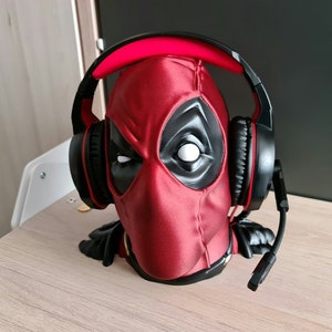 Può includere: Un supporto per cuffie a forma di testa di Deadpool rosso e nero. Il supporto indossa un auricolare da gioco nero e rosso con microfono. Il supporto è su una superficie marrone chiaro.