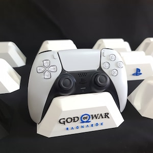 Può includere: Un controller bianco PlayStation 5 DualSense è posizionato su un supporto bianco con le parole "God of War Ragnarök" stampate sul davanti.