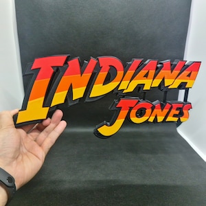 Puede incluir: Un letrero impreso en 3D con las palabras "Indiana Jones" en una fuente en negrita y estilizada. Las letras son rojas, naranjas y amarillas, con contornos negros.