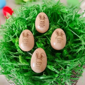 May include: Four wooden Easter eggs with bunny ears and personalized messages. The eggs are sitting on a bed of green grass. The messages on the eggs read: "Jade Happy Easter 2023", "Jimmy My First Easter 2023", "Florence Easter 2023", and "Tummy My First Easter 2023".