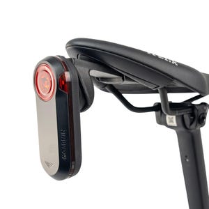 Może przedstawiać: Czarne siodełko rowerowe z czarną sztycą i czerwono-srebrną lampką tylną Garmin. Lampa ma czerwoną soczewkę, a logo Garmin jest widoczne. Na siodełku widnieje napis "FIZIK".