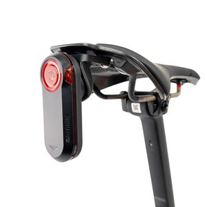 Puede incluir: Un sillín de bicicleta negro con una luz trasera Garmin roja adjunta. La luz es rectangular con una lente roja y el logotipo de Garmin. El sillín es negro y curvado. La tija del sillín es negra.