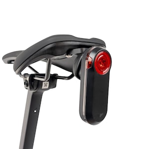 Op de afbeelding: Zwart fietszadel met een rood achterlicht bevestigd. Het licht is rechthoekig met een rode lens en is onder het zadel gemonteerd. Zadel en licht staan tegen een witte achtergrond.