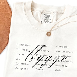 May include: A beige t-shirt with the word "Hygge" written in black cursive. The shirt has a list of words associated with the concept of hygge, including "coziness", "warmth", "intimacy", "serenity", "comfort", "contentment", "tranquility", "peaceful", "joy", "quietude", "snugness", "relaxation", "reassurance", "balance", "cheer", and "calm".
