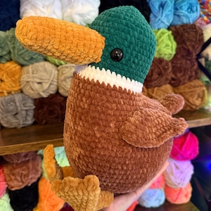 May include: A handmade crochet duck plushie with a green head, brown body, and orange beak and feet. The duck has a white collar and a black eye. The plushie is displayed in front of a shelf filled with yarn.