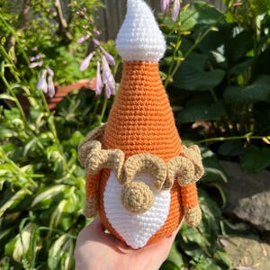 May include: A crocheted gnome with an orange body, white hat, and brown beard. The gnome is holding a white heart in its hands.