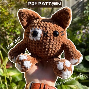 May include: A brown crocheted plush cat toy with white paw pads and a white patch over one eye. The toy features black plastic eyes and embroidered whiskers. Text on the image reads: Beans the Bean Kitty, PDF PATTERN, LOW SEW!, @krochetingkitty.