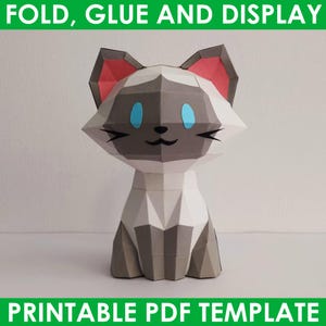 Op de afbeelding: Een printbare papercraft-sjabloon van een Siamese kat. De kat zit met zijn poten onder zijn lichaam. De kat is grijs, wit en roze met blauwe ogen. De sjabloon heeft de titel "FOLD, GLUE AND DISPLAY" en "PRINTABLE PDF TEMPLATE".
