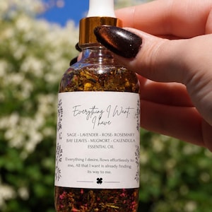 May include: A clear glass dropper bottle filled with botanical oil containing visible herbs and petals. The white label features the text Everything I Want, I have, a list of ingredients including sage, lavender, rose, rosemary, bay leaves, mugwort, and calendula, and the phrase Everything I desire, flows effortlessly to me. All that I want is already finding its way to me.