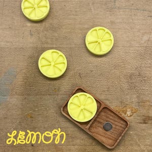 May include: Four yellow lemon-shaped coasters with a wooden tray and a small silver magnet. The text "Lemon Drop" is written in yellow letters on a wooden surface.