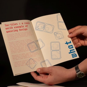 Op de afbeelding: Een open boek met de titel "Tac-tiles: a real-world example of queering design" in rode tekst. De pagina's hebben blauwe lijntekeningen en het woord "what" in blauw. Het boek wordt opengehouden door handen.