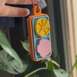 May include: A 3D-printed keychain with an orange frame, blue interior, and a yellow lemon slice and pink flower design. The keychain is attached to an orange beaded chain.
