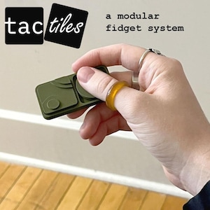 May include: An olive green modular fidget system with the text "tactiles" and "a modular fidget system". The system is held in a hand, with a yellow ring on a finger.