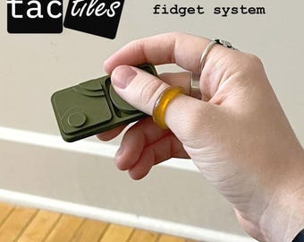 May include: An olive green modular fidget system with the text "tactiles" and "a modular fidget system". The system is held in a hand, with a yellow ring on a finger.