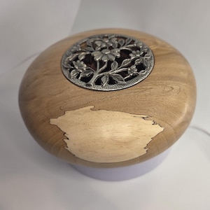 Pode incluir: Caixa decorativa de madeira com acabamento castanho claro e design floral prateado na parte superior. A caixa tem uma incrustação clara na lateral. A caixa é redonda e assenta numa base branca.