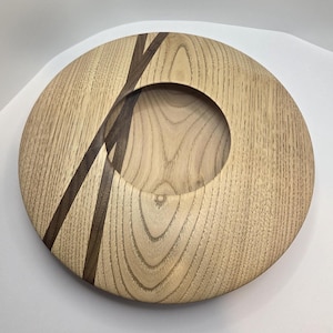 Könnte beinhalten: Eine runde Holzschale mit natürlicher Holzmaserung. Die Schale hat eine kreisförmige Öffnung in der Mitte und ist mit zwei dunkelbraunen, diagonalen Streifen akzentuiert. Die Schale wird wahrscheinlich zu dekorativen Zwecken verwendet.