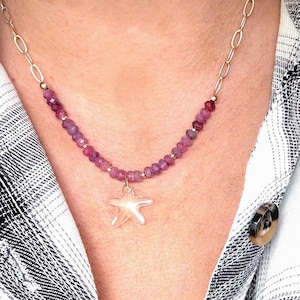 May include: A gold chain necklace with a strand of small, pinkish-purple beads. A silver starfish pendant hangs from the center. The necklace is worn over a black and white plaid shirt.