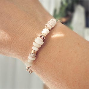 May include: A delicate bracelet featuring white shell beads and gold-colored accents. The bracelet is strung together with a fine chain and clasp. The bracelet is worn on a wrist.