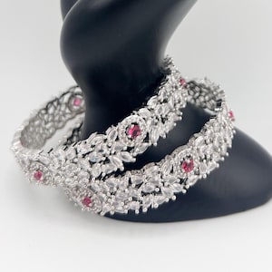 May include: Two silver bangles with white stones and pink accents. The bangles are displayed on a black hand-shaped stand.