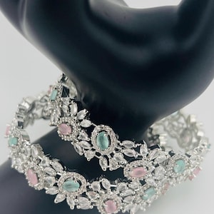 May include: Two silver bangles with white and clear stones, accented with pink and light blue oval-shaped stones.