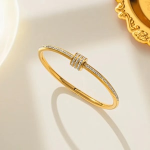 May include: A gold-toned bangle bracelet featuring a central decorative element composed of three stacked rings encrusted with small clear stones. The band of the bracelet is also inlaid with a continuous row of clear stones along its top surface.