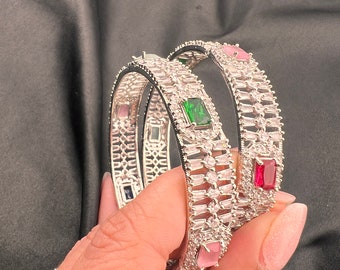 May include: Two silver bangles with clear and coloured stones. The bangles are decorated with rows of clear stones and feature a pink, green, and red stone on each bangle.