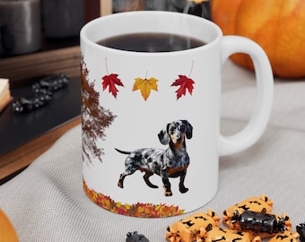 Op de afbeelding: Witte keramische koffiemok met een illustratie van een zwart-witte teckel, herfstbladeren en een bruine boom. De mok is gevuld met koffie en heeft een zwart handvat.