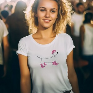 Op de afbeelding: Een vrouw met blond krullend haar draagt een wit T-shirt met een roze teckel die een roze hoed en roze laarzen draagt. Ze glimlacht naar de camera.