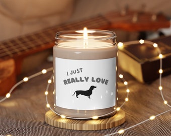 May include: A brown candle with a white label that says "I just really love" with a silhouette of a dachshund. The candle is on a wooden coaster and surrounded by white fairy lights.